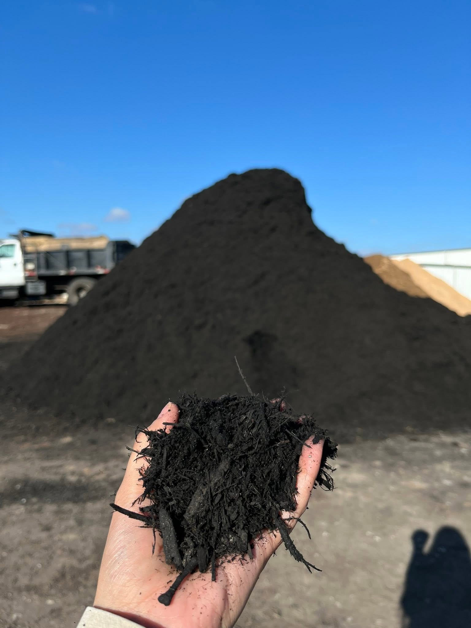 Dark mulch shown by hand in front of a stockpile
